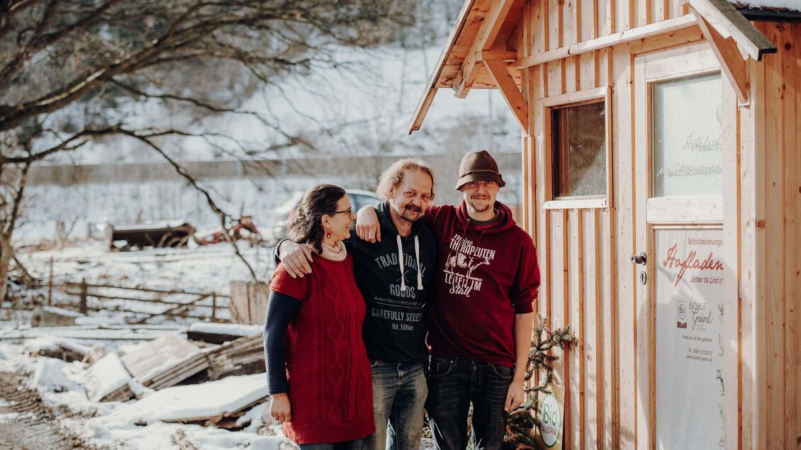 Drei Personen stehen lächelnd vor einem kleinen Holzhaus. Die Umgebung ist winterlich mit Schnee und einer ruhigen Landschaft. | © GenussReich
