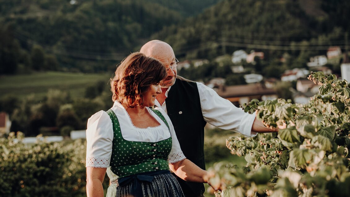 Ein Paar in traditioneller Kleidung steht in einem Weinberg und betrachtet die Pflanzen. Im Hintergrund sind sanfte Hügel und ein paar Häuser sichtbar. | © Freilichtmomente