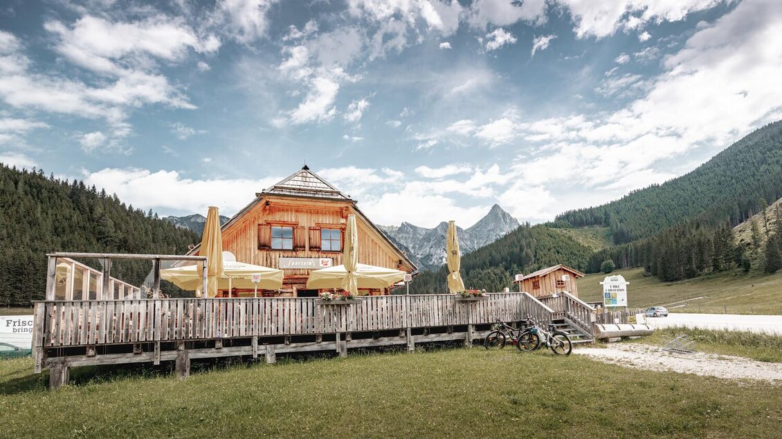 Ein altes Holzhaus mit einer großzügigen Terrasse steht inmitten einer grünen Wiese. Im Hintergrund erstrecken sich majestätische Berge unter einem bewölkten Himmel. | © Thomas Sattler