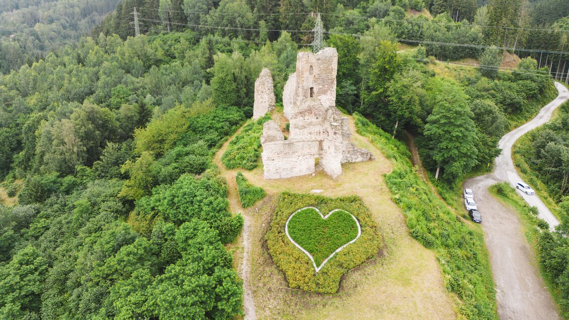 Eine alte Burgruine steht auf einem Hügel, umgeben von üppigem Grün. Im Vordergrund ist ein Herz aus Blumen zu sehen. | © Karl Steinegger