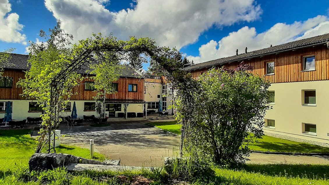 <p>Die Gebäude des JUFA Hotels mit Holzverkleidung und großen Fenstern.Im grünen Garten gibt es hohe Sträuche und einen Bogen. Im Hintergrund befindet sich eine Terrasse mit Tischen und Sonnenschirmen.</p> | © JUFA Hotels