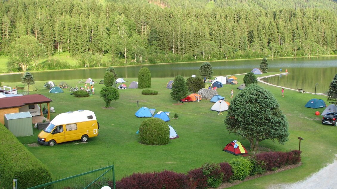 Ein Campingplatz am See, umgeben von Bäumen und Bergen. Verschiedene Zelte und ein Wohnmobil sind auf dem Grün verteilt. | © Hannes Zenz