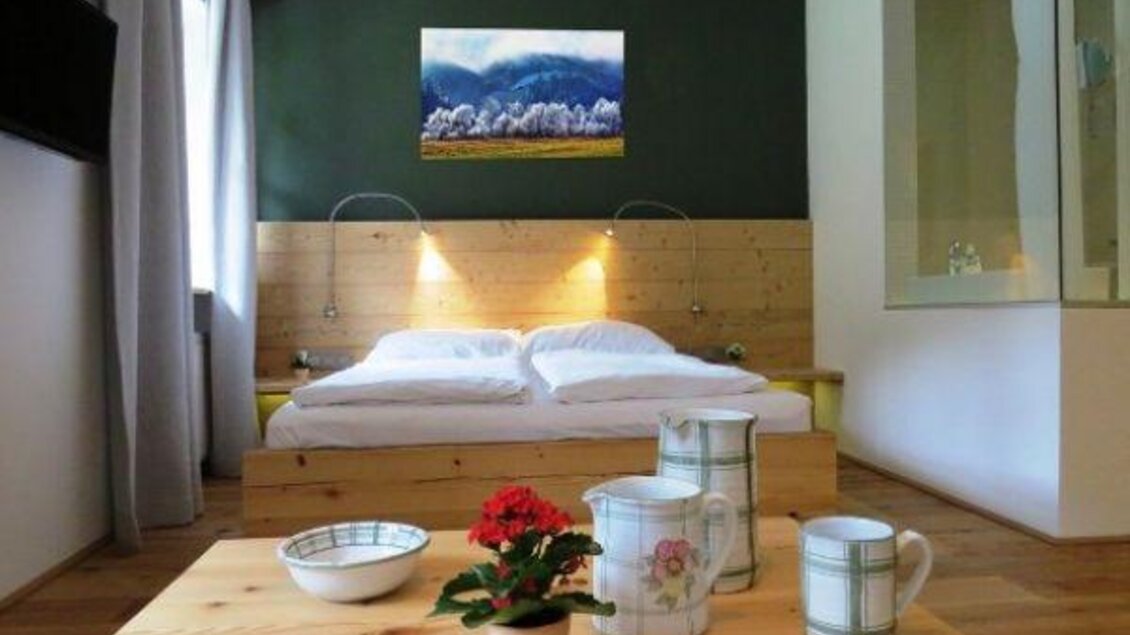 Ein gemütliches Schlafzimmer mit einem Holzbett und einem modernen Wandgemälde. Auf dem Tisch stehen eine Blume, eine Schüssel und zwei Kannen. | © ZWHS
