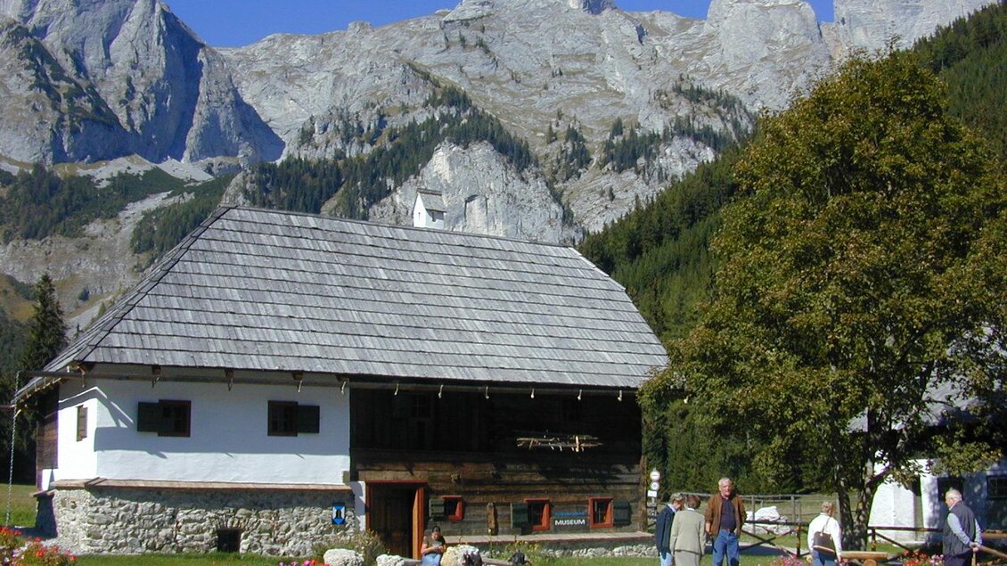 Ein traditionelles Holzhaus vor einer beeindruckenden Berglandschaft. Menschen stehen im Garten und genießen die Umgebung. | © ZWHS