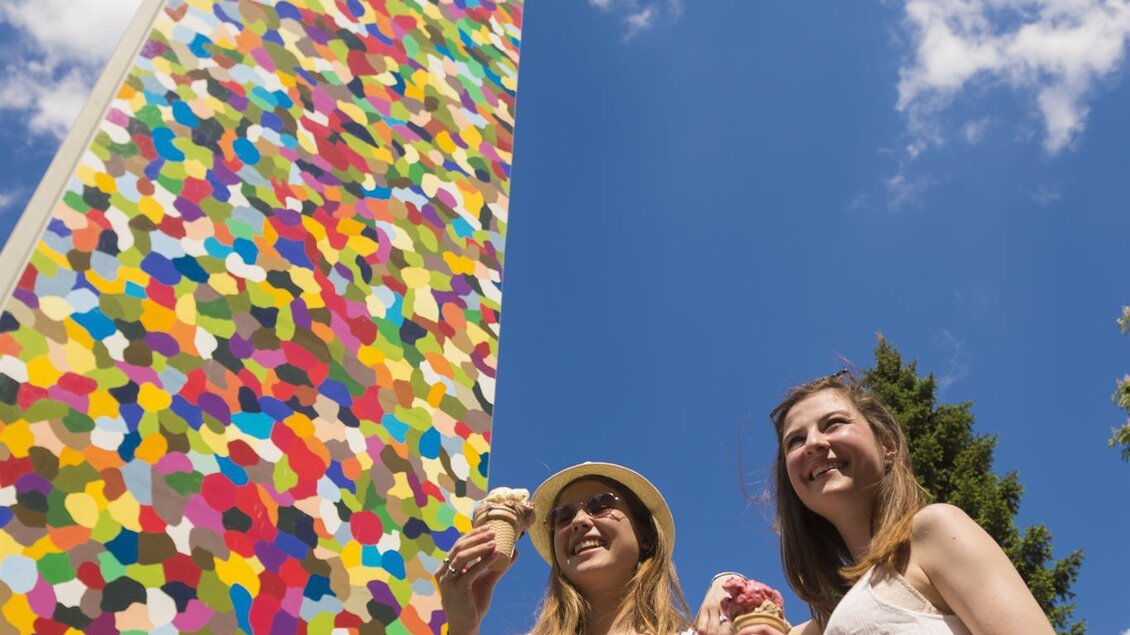 Zwei Frauen stehen vor einem bunten, hohen Turm und genießen Eis. Der Himmel ist blau mit einigen Wolken. | © Tourismusverband Feldbach/ B. Bergmann