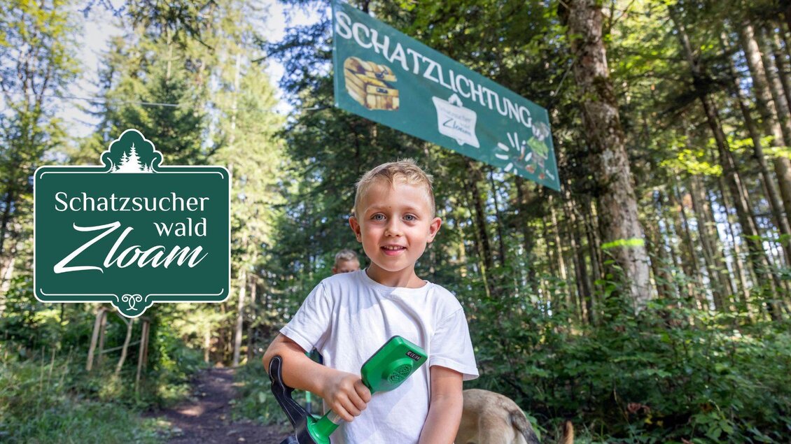 Ein lächelnder Junge hält eine Schatzsuche-Ausrüstung in einem Wald. Im Hintergrund ist ein Schild mit der Aufschrift "Schatzlichtung" zu sehen. | © www.zloam.at