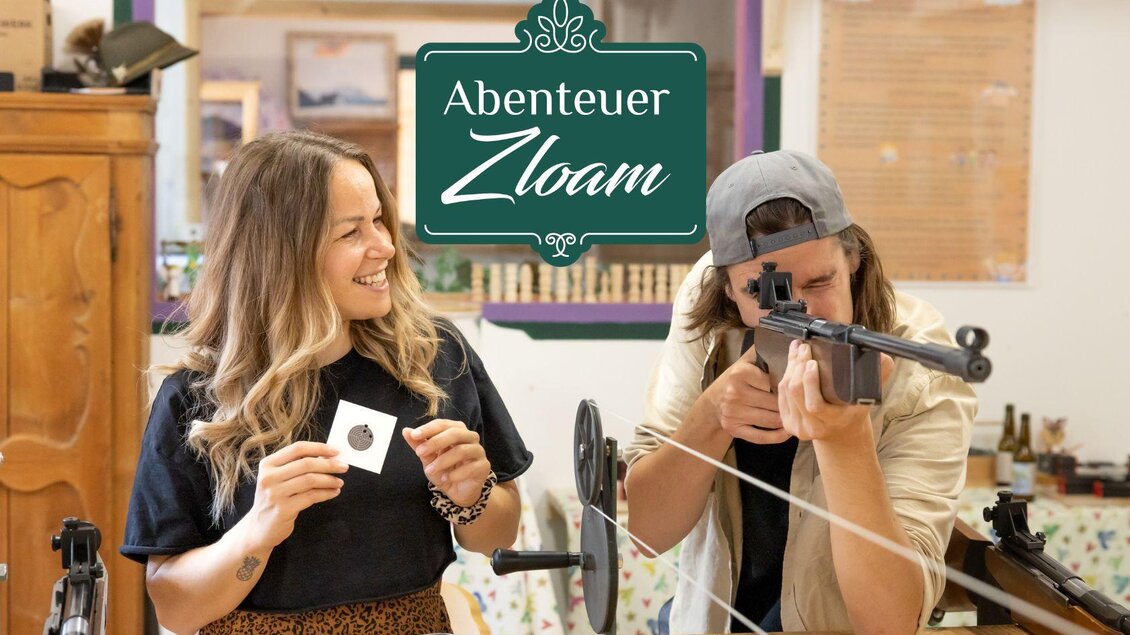 Eine lächelnde Frau zeigt einen Gegenstand, während ein Mann mit einem Gewehr auf ein Ziel zielt. Im Hintergrund ist der Schriftzug "Abenteuer Zloam" zu sehen. | © www.zloam.at