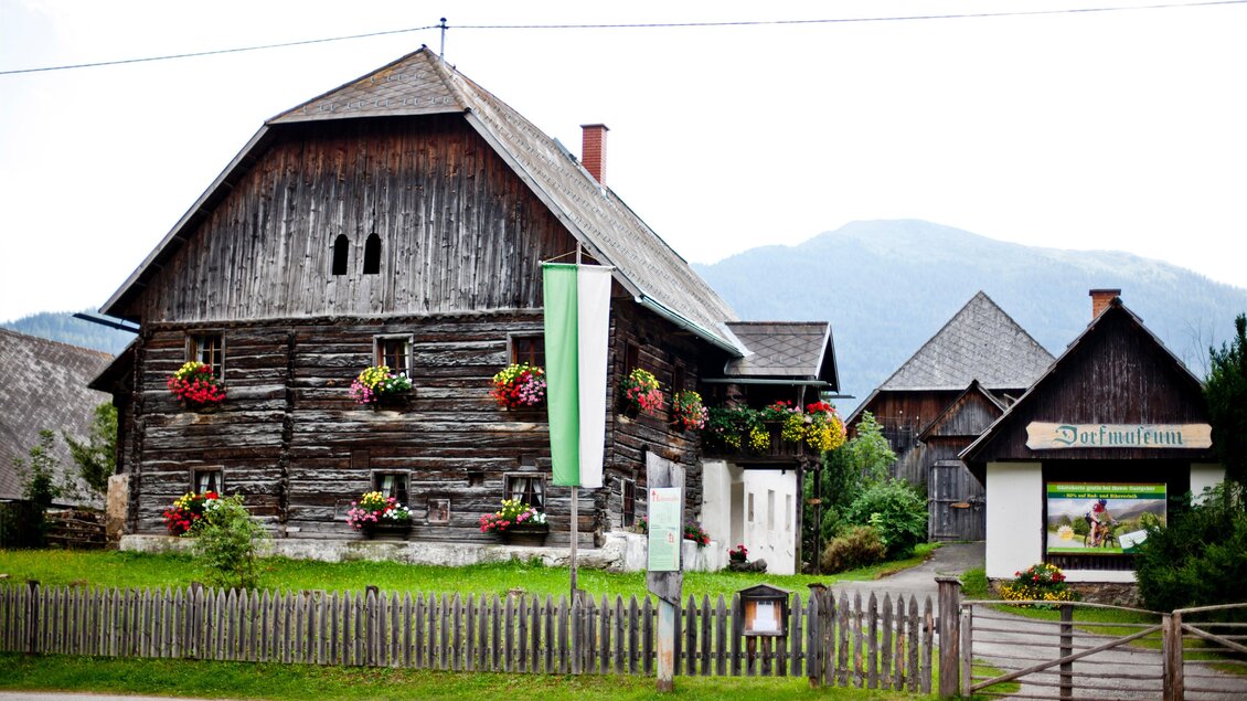 Ein traditionelles Holzhaus mit blühenden Blumenkästen. Im Hintergrund sind Berge und eine ruhige Landschaft zu sehen. | © Holzwelt Murau