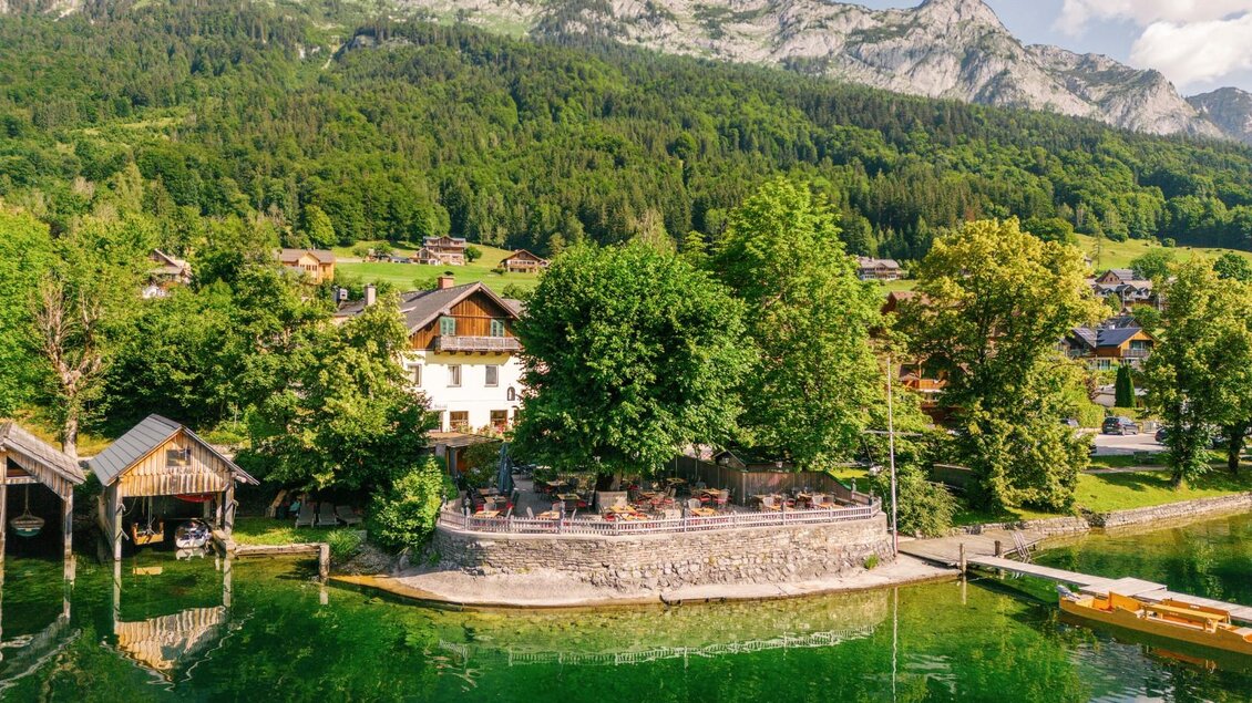 Ein ruhiger See umgeben von Bergen und üppigem Grün. Im Vordergrund sind Bäume und ein Haus mit einer Terrasse zu sehen. | © Gasthaus Stöckl
