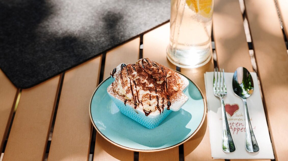 Ein Teller mit einer leckeren Nachspeise, dekoriert mit Schokolade. Neben dem Dessert steht ein Glas Wasser mit Zitrone und Besteck auf einem Holztablett. | © Gasthaus Stöckl