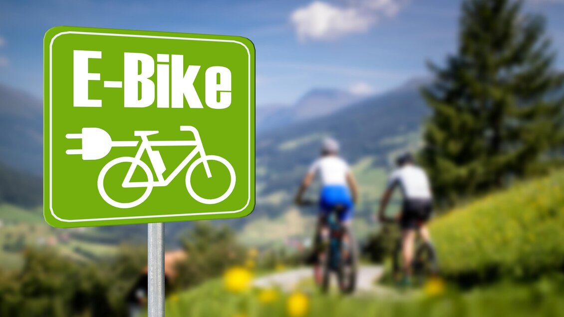 Ein Schild mit der Aufschrift "E-Bike" zeigt, dass hier E-Bikes genutzt werden können. Im Hintergrund sind Radfahrer in einer malerischen Landschaft sichtbar. | © TV Murau