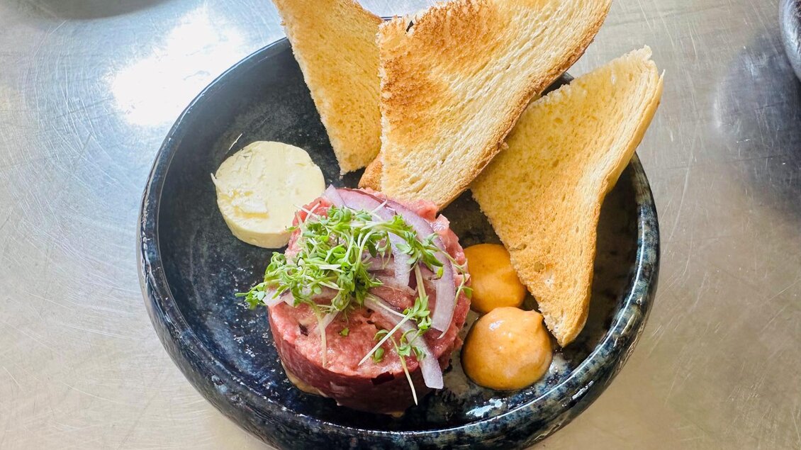 Ein Teller mit einer Fleischscheibe, garniert mit Zwiebeln und frischen Kräutern. Dazu goldene Toaststücke und zwei Saucen in kleinen Schalen. | © Eagle - Wirtshaus am Golfplatz