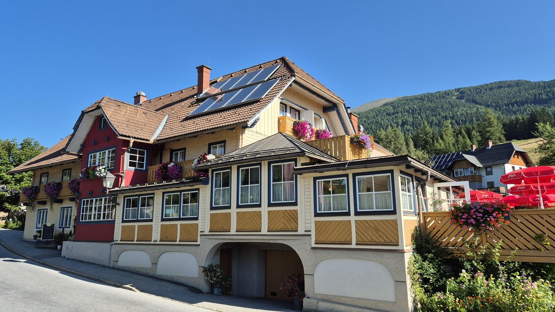 Ein schönes Mehrfamilienhaus mit bunten Blumenkästen und Solarpanelen. Die Umgebung ist von grünen Bergen und klarem Himmel geprägt. | © TVB Murau