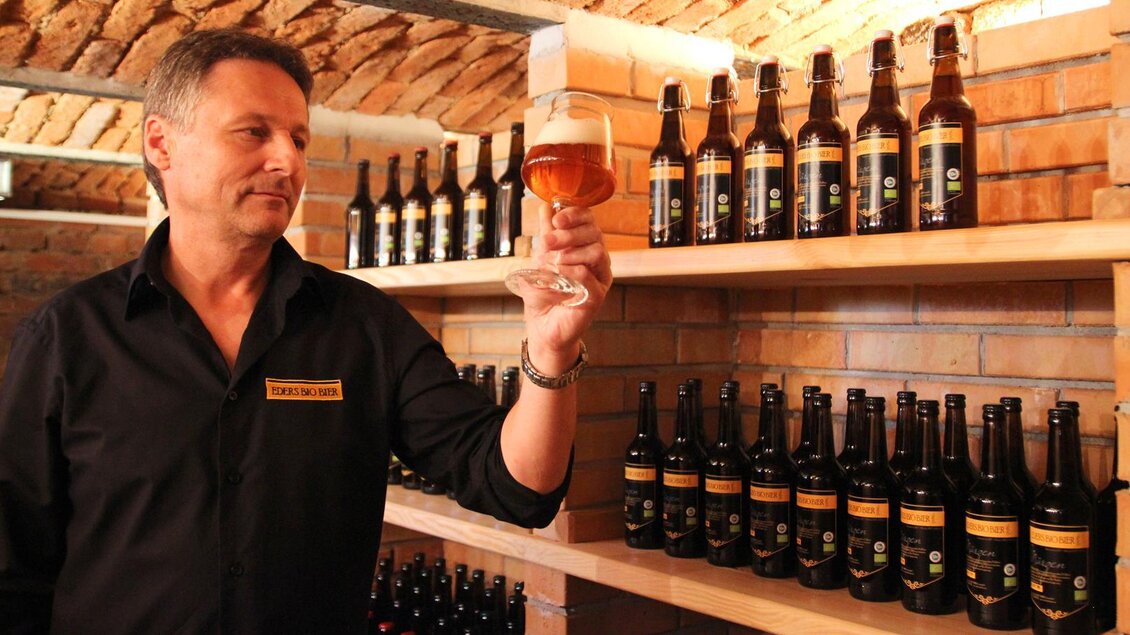 Ein Mann in einem schwarzen Hemd hält ein Glas Bier und betrachtet es kritisch. Im Hintergrund stehen zahlreiche Bierflaschen in einem Regal. | © Eders Bio Bier