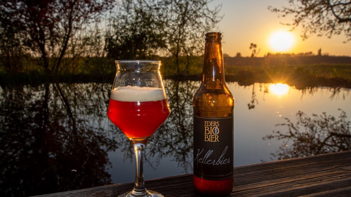 Ein Glas rotes Bier steht neben einer Flasche auf einem Tisch. Im Hintergrund ist ein schöner Sonnenuntergang über einem ruhigen Gewässer zu sehen. | © Eders Bio Bier