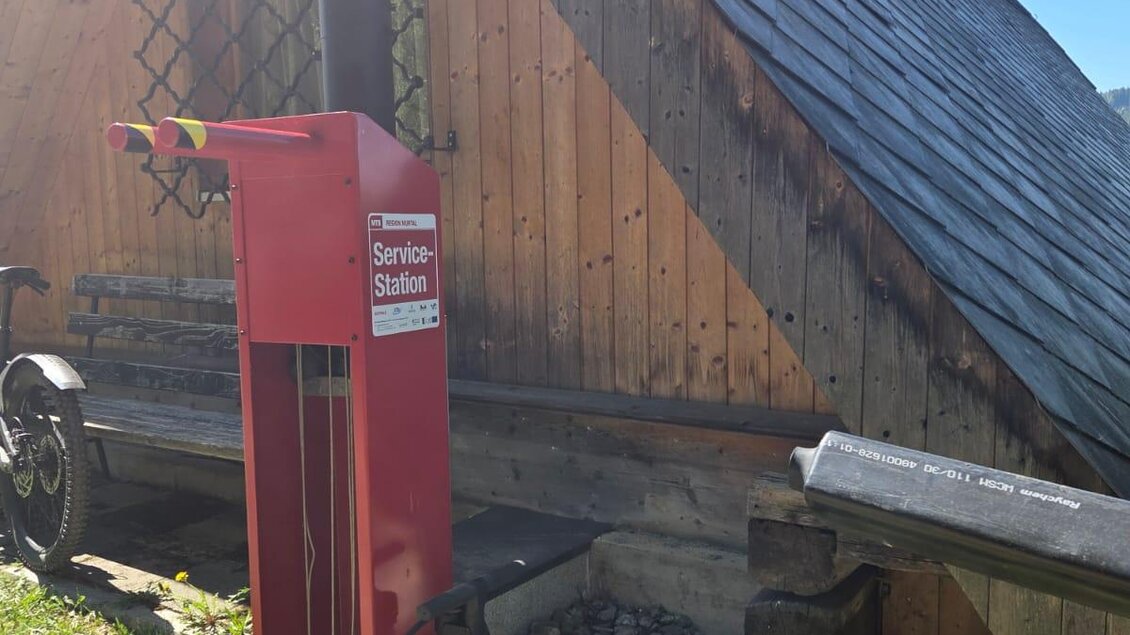 Eine rote Servicestation steht neben einem Holzhaus. Im Hintergrund liegt eine grüne Wiese und ein blauer Himmel. | © Roman Gruber