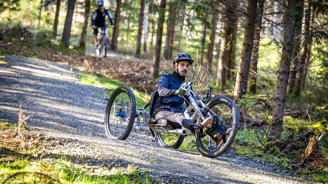 Ein Mann fährt mit einem Handbike auf einem schmalen Waldweg. Im Hintergrund ist ein weiterer Radfahrer sichtbar. | © Christian Webersink