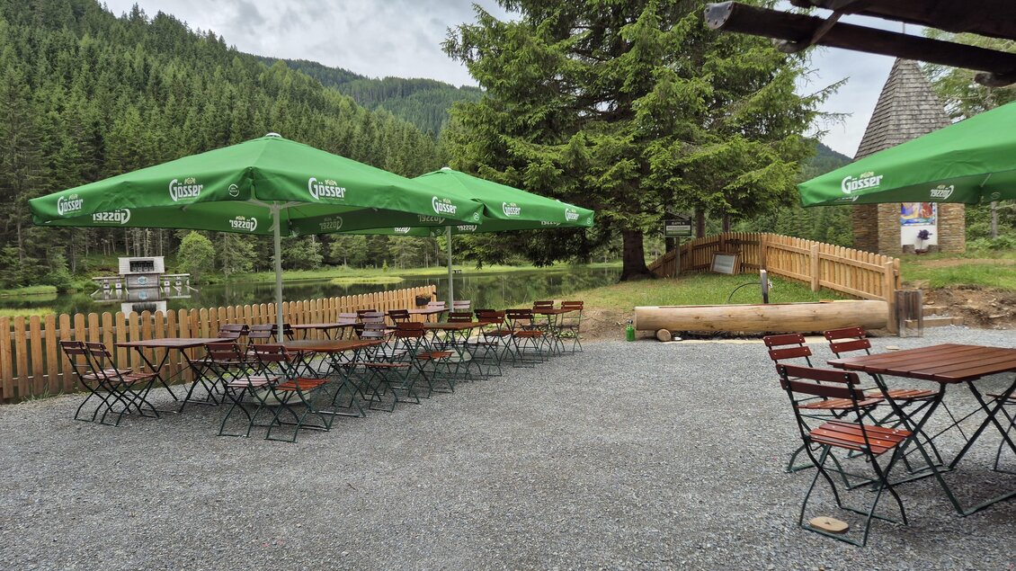 Eine kleine Terrasse mit Tischen und Stühlen unter grünen Schirmen. Im Hintergrund sind Bäume und eine Berglandschaft zu sehen. | © TVB Murau