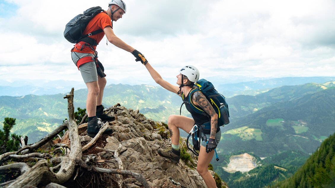 Zwei Kletterer unterstützen sich gegenseitig beim Besteigen eines felsigen Gipfels. Im Hintergrund sind grüne Hügel und ein bewölkter Himmel zu sehen. | © Oststeiermark Tourismus