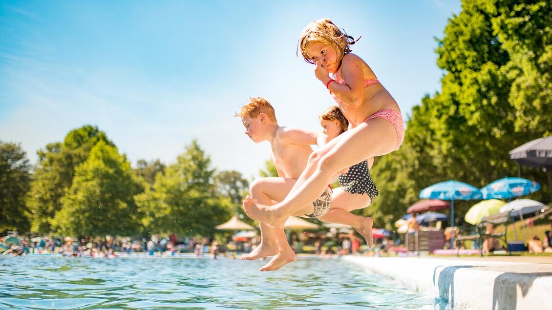 Drei Kinder springen fröhlich ins Wasser. Im Hintergrund sind Sonnenliegen und Bäume zu sehen. | © Tourismusverband Thermen- & Vulkanland