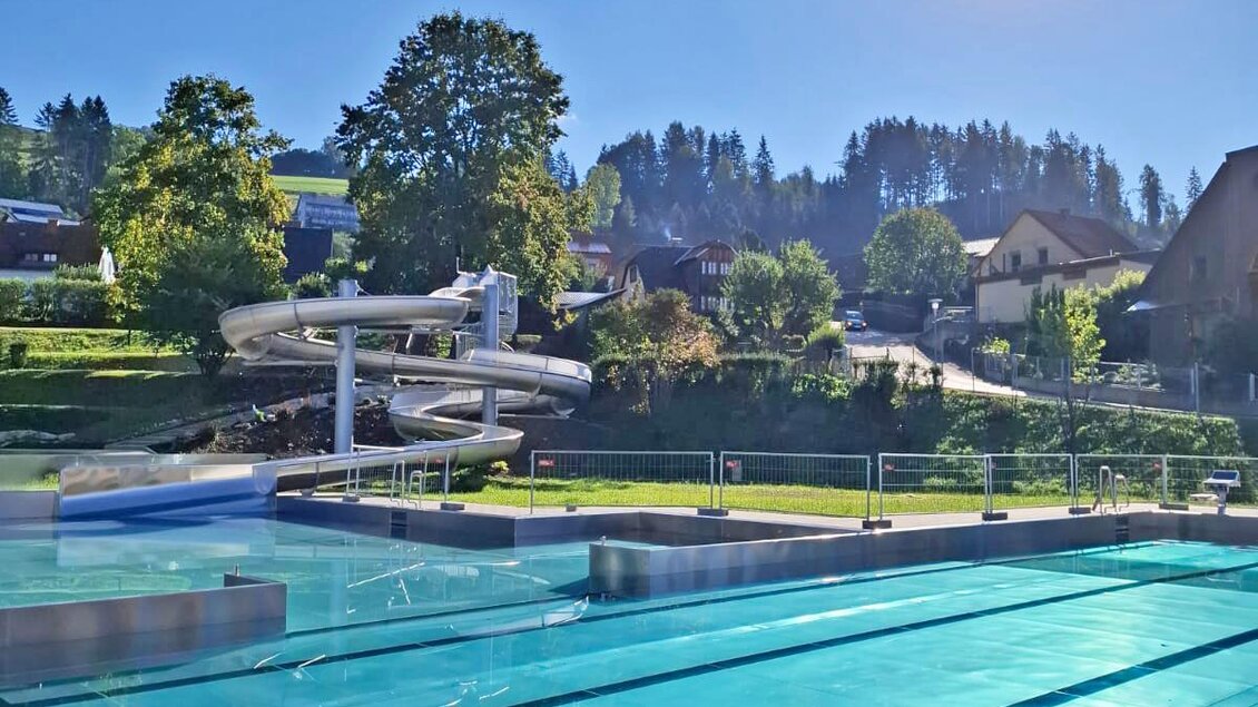 Ein Schwimmbad mit klarem Wasser und einem Wasserrutschen. Im Hintergrund sind Bäume und Häuser vor einem blauen Himmel zu sehen. | © Gemeinde Neumarkt in der Steiermark