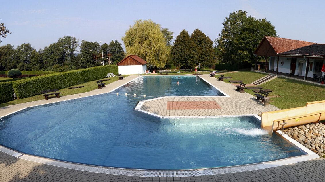 Ein großer Swimmingpool mit klarem Wasser und einer Wasserrutsche. Umgeben von einer grünen Wiese und Bäumen gibt es Sitzgelegenheiten und ein Gebäude im Hintergrund. | © Stadtgemeinde Fürstenfeld