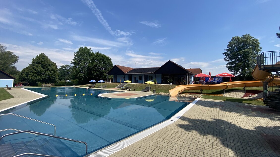 Ein Schwimmbad mit klarem Wasser und einer Rutsche. Im Hintergrund sind grüne Bäume und Sitzmöglichkeiten unter bunten Sonnenschirmen. | © Stadtgemeinde Fürstenfeld