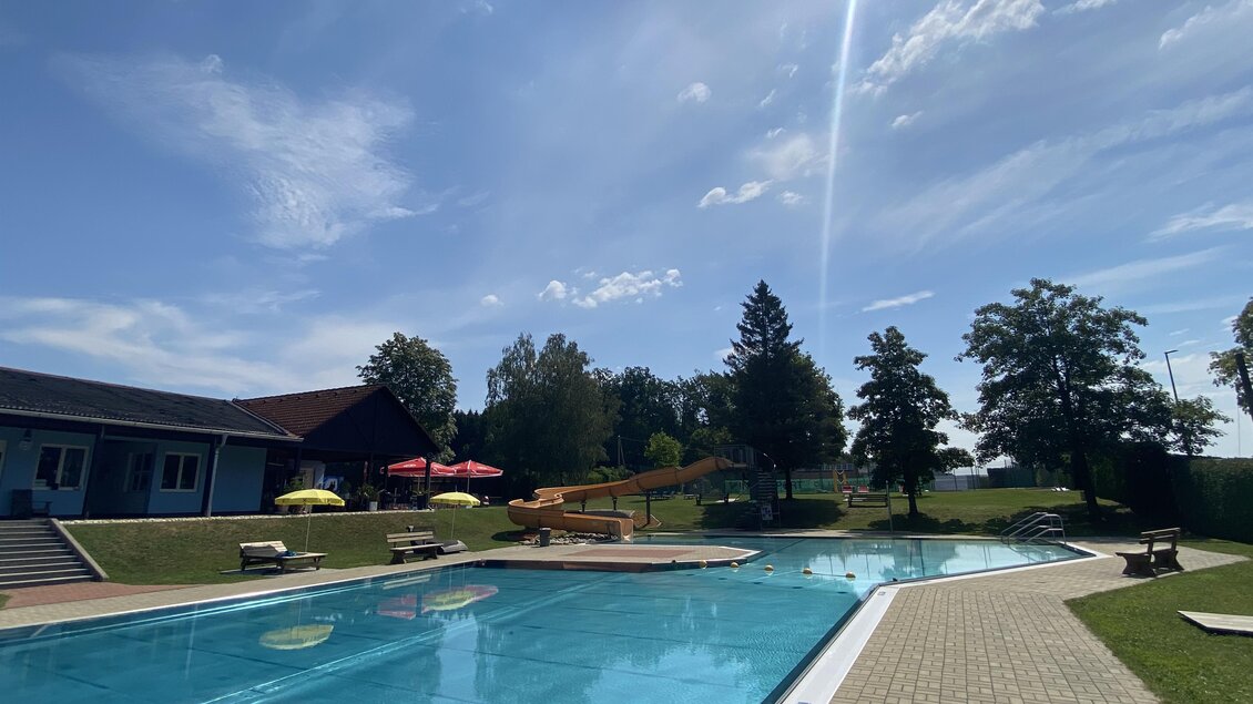 Ein Schwimmbecken an einem sonnigen Tag mit blauen Himmel und wenigen Wolken. Im Hintergrund sind Liegestühle und parasolartige Schirme sichtbar. | © Stadtgemeinde Fürstenfeld