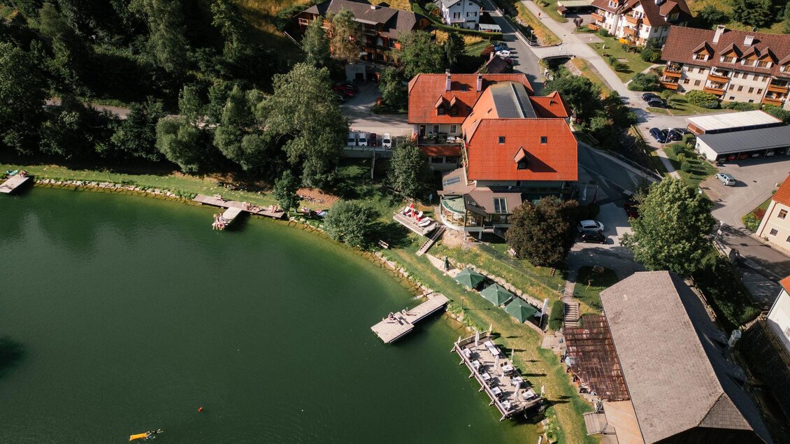 Ein malerisches Gebäude am Ufer eines ruhigen Sees, umgeben von grünen Hügeln. Das Wasser spiegelt die bunten Häuser und die Landschaft wider. | © Christoph Lukas