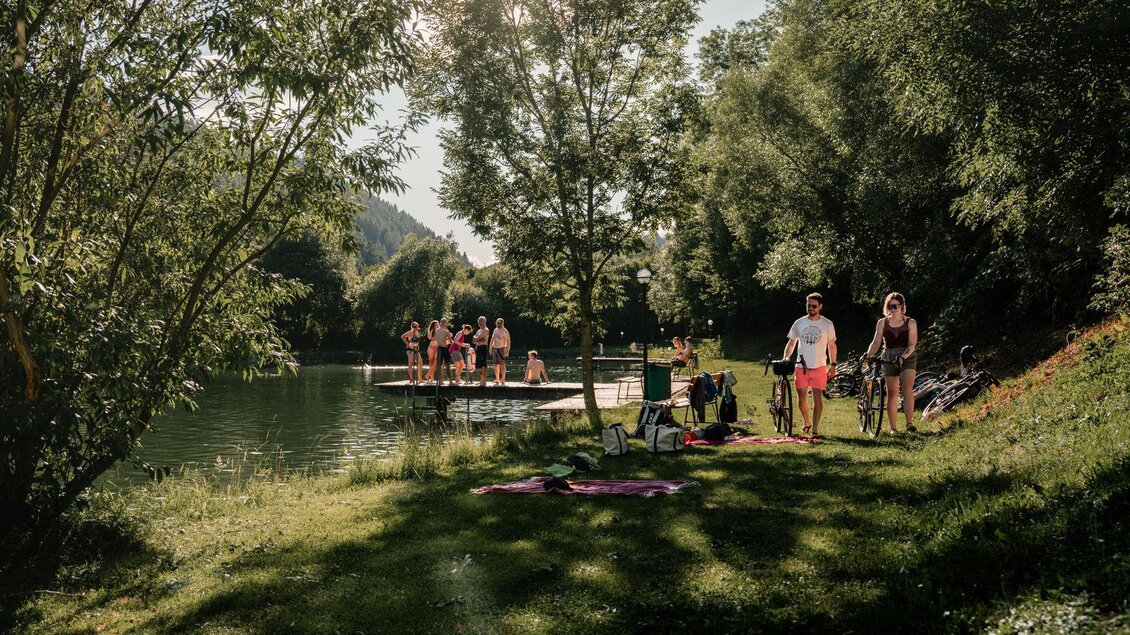 <p>Ein ruhiger Teich mit grünem Wasser, umgeben von Bäumen und Gras. Die Sonne scheint auf die Wasseroberfläche und reflektiert die Umgebung. Im Bild sind Badegäste und Radfahrer zu sehen.</p> | © Christoph Lukas