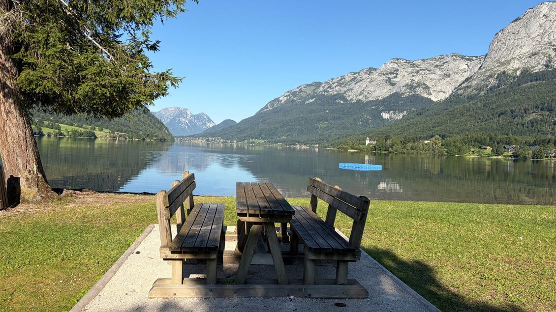<p>Eine Holzbank mit Tisch steht am Ufer des Grundlsees beim Freizeitzentrum in Gößl. Am See schwimmt auch ein blaues Floß.</p> | © TVB Ausseerland/Johanna Rastl
