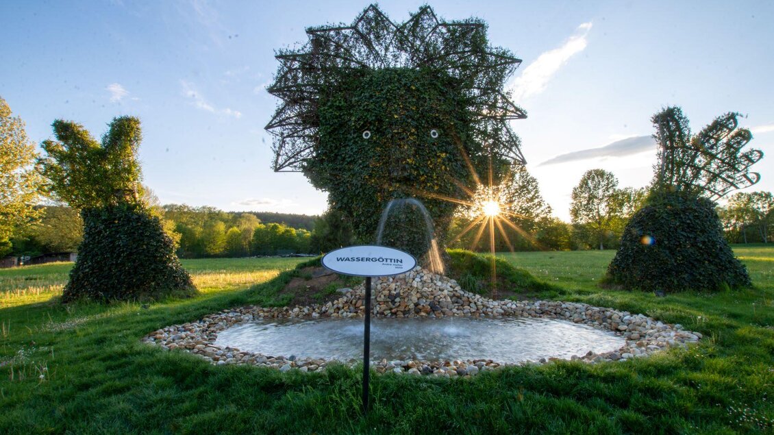 Eine kreative Gartenlandschaft mit lebenden Pflanzen in Form von Tieren. Im Hintergrund scheint die Sonne durch die Bäume und reflektiert auf dem Wasserbecken. | © Kurkommission Bad Blumau