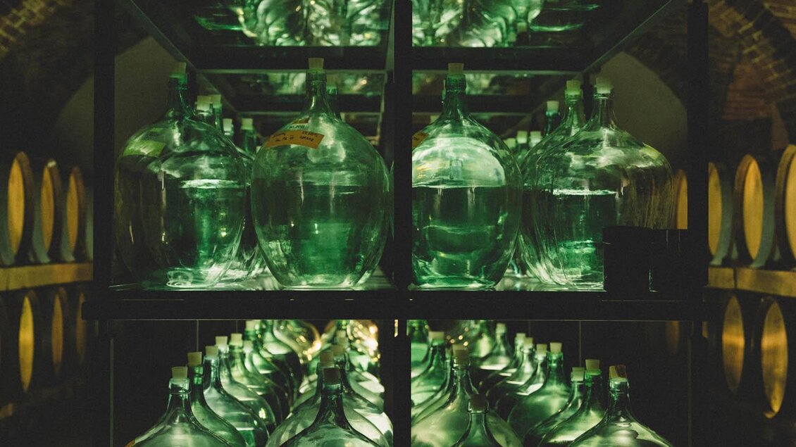 Eine Reihe grüner Glasbehälter steht auf Regalen in einem Keller. Die Behälter werden von einer sanften Beleuchtung erhellt. | © Manufaktur Gölles