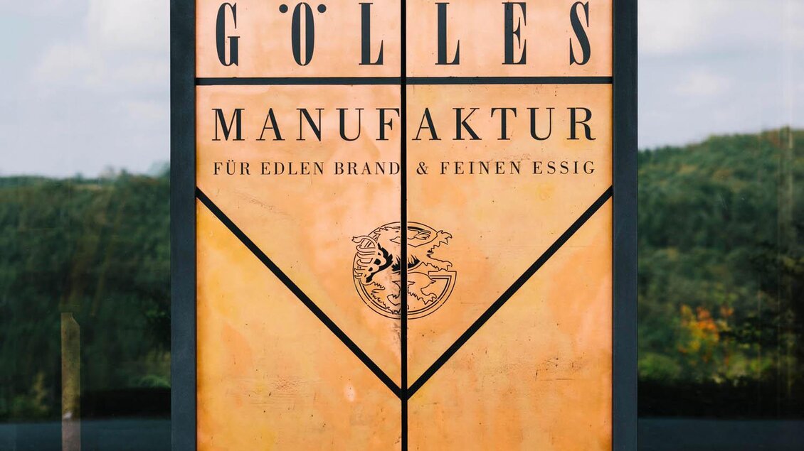 Eine Tür mit der Aufschrift "GÖLLES MANUFAKTUR" für edlen Brand und feinen Essig. Der Hintergrund zeigt eine grüne, bewaldete Landschaft. | © Manufaktur Gölles