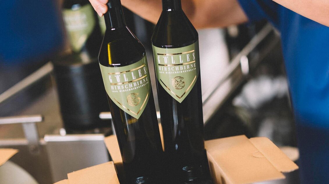 Eine Person packt zwei Flaschen Wein in einen Karton. Im Hintergrund sind weitere Flaschen zu sehen. | © Manufaktur Gölles