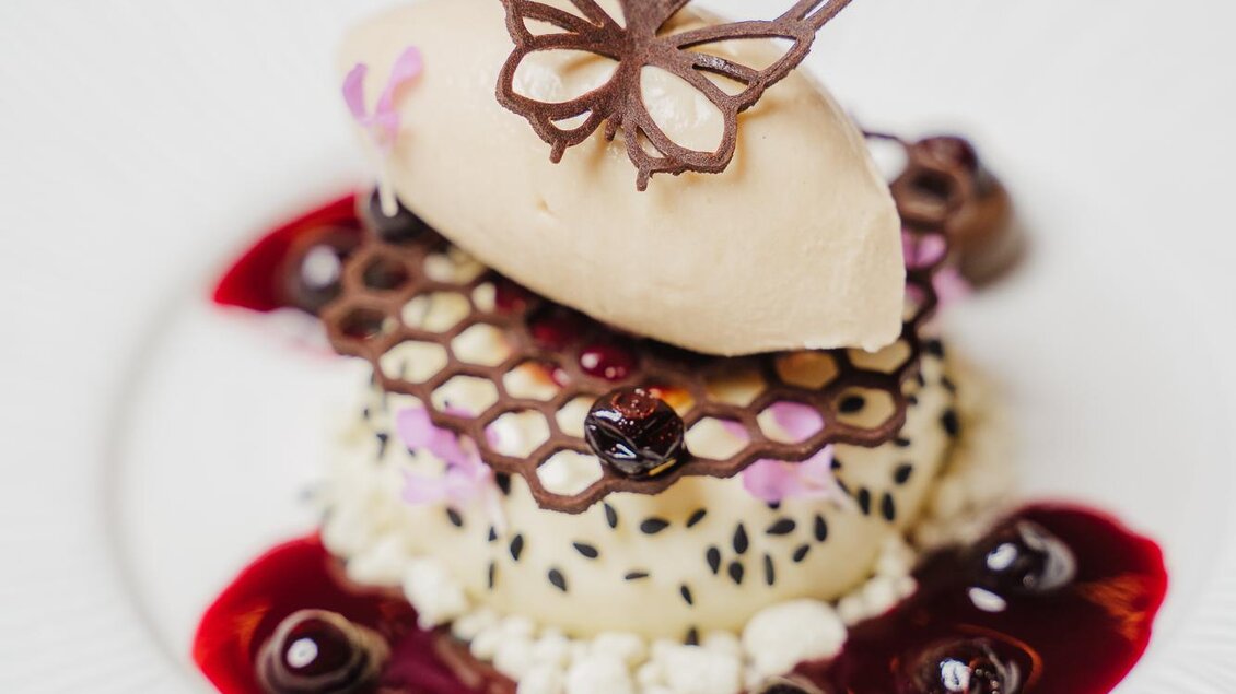Ein kunstvoll angerichtetes Dessert mit einer Mousse und einer Schokoladenbutterfliege. Das Gericht ist mit einer fruchtigen Soße und bunten Dekoren verziert.