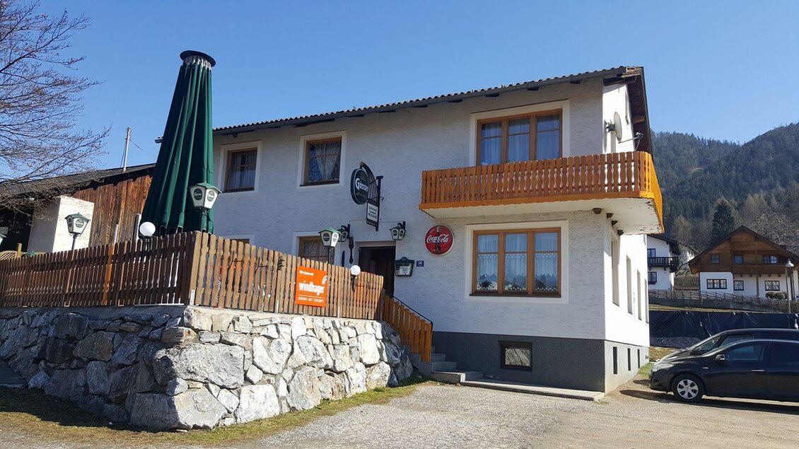 Ein Gasthaus mit einem Erdgeschoss und einem Balkon. Im Hintergrund sind Berge und ein blauer Himmel sichtbar. | © Gasthaus Mossauer