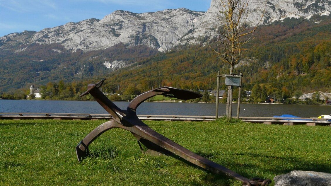 Ein großer Anker liegt auf der Wiese am Seeufer. Im Hintergrund sind majestätische Berge und Bäume zu sehen. | © TVB Ausseerland - Salzkammergut