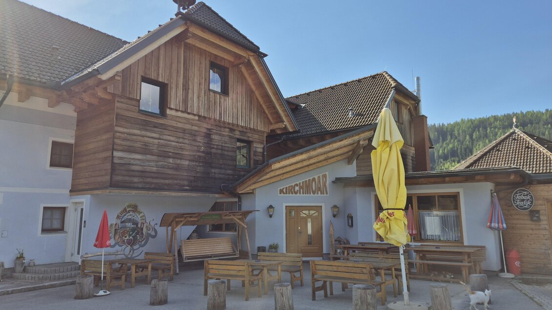 Ein rustikales Gasthaus mit Holzfassade und einer einladenden Terrasse. Im Vordergrund stehen mehrere Holztische und -bänke, umgeben von einer malerischen Landschaft. | © TV Murau