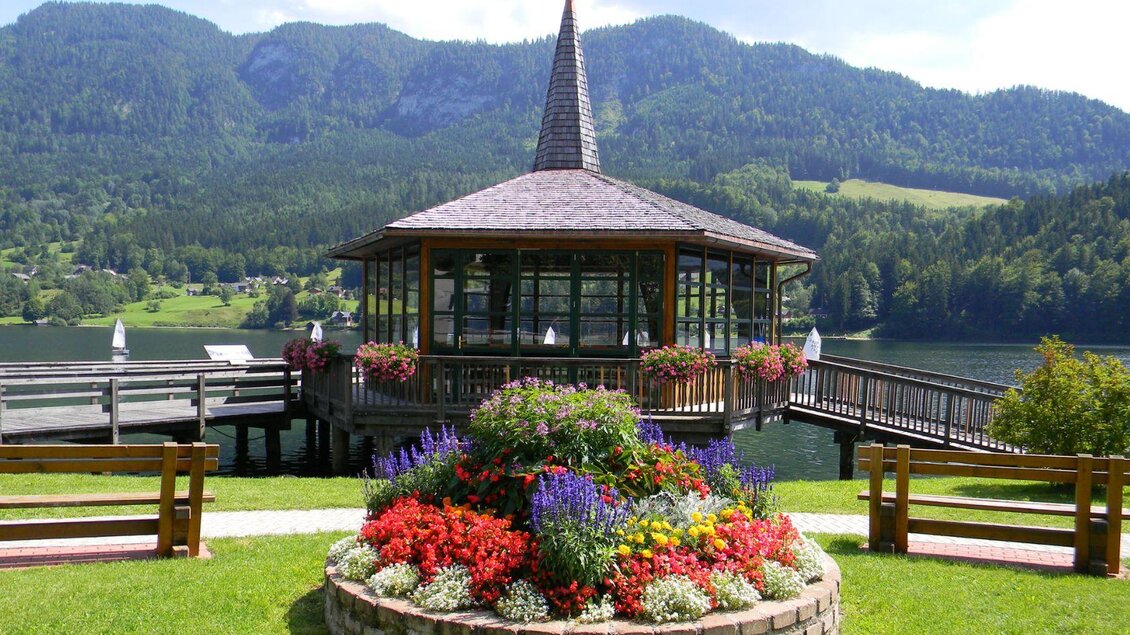 Ein schöner Pavillon am Wasser, umgeben von bunten Blumenbeeten. Im Hintergrund sind grüne Berge und ein klarer, blauer Himmel zu sehen. | © Gemeinde Grundlsee