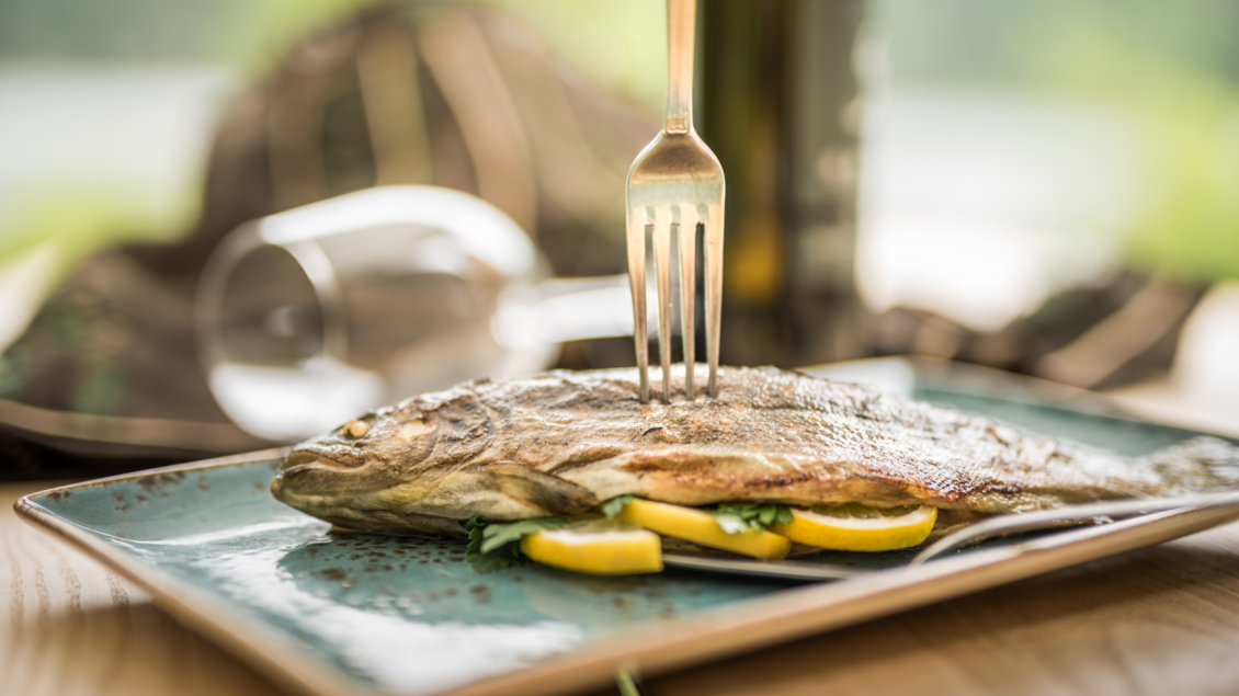 <p>Ein frisch zubereiteter Saibling, ein hier geläufiger Fisch aus den Ausseerland Gewässern, der auf einem Teller mit Zitronenscheiben garniert angerichtet ist. Im Hintergrund sind ein Glas und eine Flasche Olivenöl zu sehen.</p> | © Genuss am See