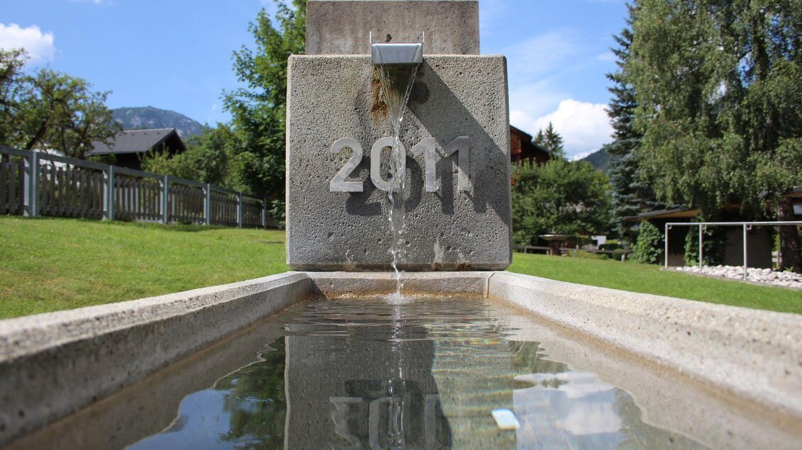 Eine moderne Steinquelle mit der Jahreszahl 2011. Klar Wasser fließt in ein langes, schmalen Becken im Freien. | © Viola Lechner