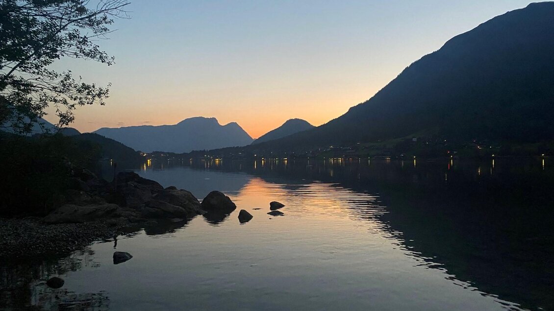Ein ruhiger See bei Sonnenuntergang, umgeben von Bergen. Das Wasser spiegelt die sanften Farben des Himmels wider. | © Bettina Scheck