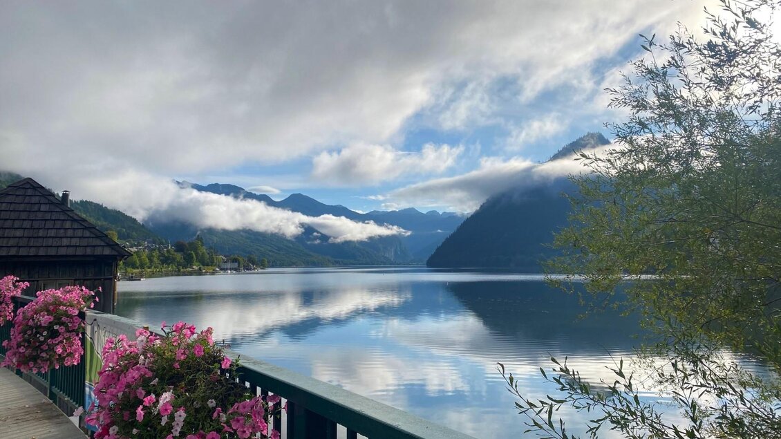 Der ruhige Grundlsee, aufgenommen beim Fischkalter am Anfang des Sees. Umgeben von den Ausläufern des Toten Gebirges ist er von sanften Wolken durchzogen. Bunte Blumen schmücken den Teil des Promenadenwegs beim Fischkalter. | © Bettina Scheck