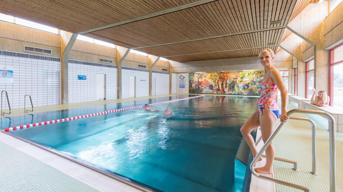Ein Innenpool mit klarem Wasser und bunten Wänden. Eine Person steht an der Seite des Pools und schaut zur Kamera. | © Stadtgemeinde Trofaiach