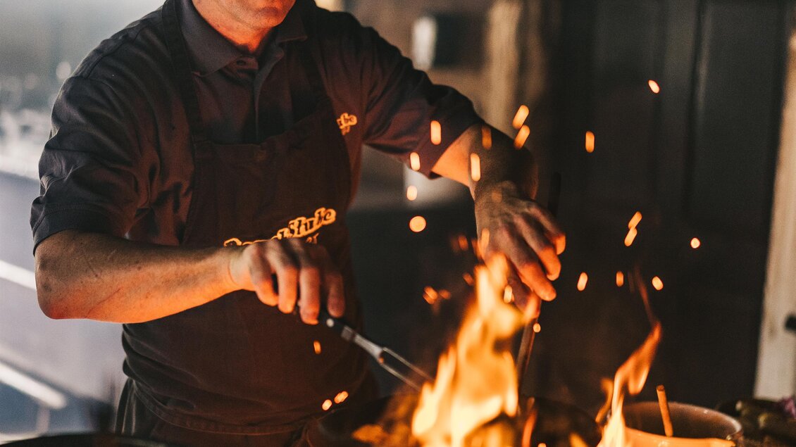 Ein Koch steht vor einem großen Feuer und rührt darin. Funken fliegen auf, während er die Speisen zubereitet. | © Oststeiermark Tourismus
