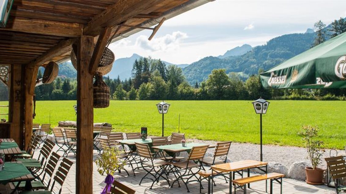 Ein einladendes Restaurant mit Holzterrasse und Tischen im Freien. Im Hintergrund ist eine malerische Landschaft mit grünen Wiesen und Bergen zu sehen.