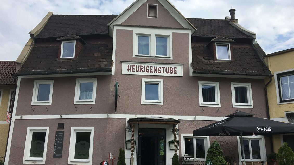 Ein Gebäude mit dem Schild „Heurigenstube“. Die Fassade ist in sanften Farben gehalten und es gibt einen Außenbereich mit Tischen. | © Familie Angerer