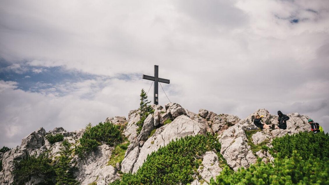 Eine Person mit einem Rucksack steht auf einem Berg und blickt auf ein Kreuz. Der Himmel ist blau mit einigen Wolken und die Umgebung ist felsig und grün. | © Oststeiermark Tourismus