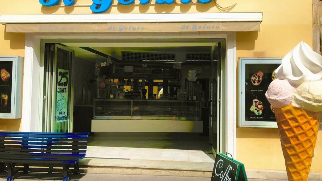 Ein Eisgeschäft mit dem Namen "Il Gelato" und einer bunten Eiscreme-Skulptur vor dem Eingang. Es gibt eine blaue Bank und ein Schild mit der Aufschrift "Coffee to go".